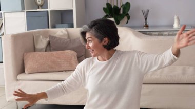 Caucasian woman, soft gray hair, dressed in white, joyfully extending arms in light-filled room, radiating peaceful energy, enjoying calming moment, sitting in stylish minimalist home interior. clipart