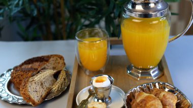 Kahvaltı sunumunda altın kruvasanlar, süslü tutacakta rafadan yumurta, otlarla dilimlenmiş çoklu tahıllı ekmek, canlı portakal suyu ve rafine edilmiş minimalist ayrıntılar yer alıyor..