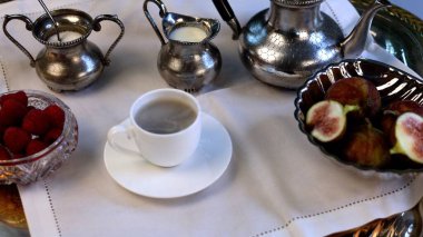 Kahvaltı masasında siyah saplı klasik gümüş çaydanlık, şeker krikosu, süt kristalleri, kahve dolu porselen bardak, kristal ahududu kasesi ve metal tepside dilimlenmiş incir bulunuyor..