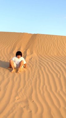 Smiling child sitting on golden sand dunes under clear blue sky. Sunlight creates soft shadows on rippling sand patterns, highlighting playful moment in serene desert landscape.  clipart