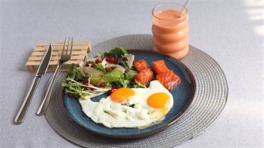 Somon dilimlerinin yanında iki güneşli omlet, nar tohumlu yeşil salata ve turuncu meyve suyu. Temiz yeme ve sağlığa odaklı dengeli çevre dostu yemek..