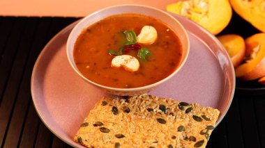 Canlı turuncu tonlarda kabak çorbası yumuşak pembe tabakta servis edilir, kıtır kıtır tohum krakerleriyle tamamlanır, arka planda sıra balkabakları, sıcaklık, temiz beslenme ve ekolojik yaşam tarzı eklenir..