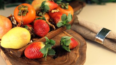 Fruit arrangement on wooden tray features fresh persimmons, strawberries, pears, blending vibrant fruit colors with natural wooden tray textures for rustic, inviting presentation.  clipart