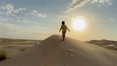 Desert sand leads child toward sun, footprints follow soft curves of golden dunes, warm glow covers landscape under fading light, peaceful journey unfolds, desert harmony concept. clipart