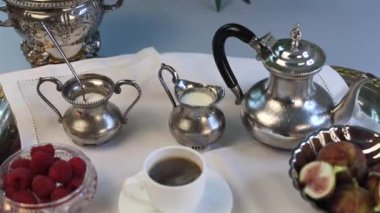Kahvaltı seti süslenmiş gümüş çaydanlık, kavisli şeker ve süt kristalleri, gümüş tepside dinlenen incirler, kristal kasedeki ahududular ve beyaz porselen bardaklar içerir.. 