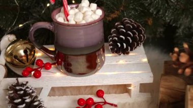 Yeşil çam ağaçları, kırmızı böğürtlen, altın çan ve çam kozalakları arasına yerleştirilmiş sıcak çikolata, marşmelov ve şeker kamışlarıyla dolu tatil kupası..