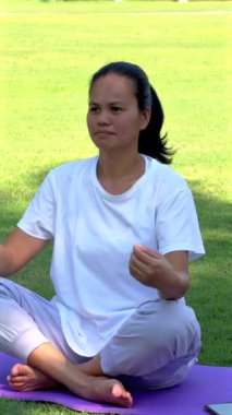 Asian woman connecting with nature, meditating on grass, dressed in white symbolizing simplicity, representing holistic well-being, natural harmony.