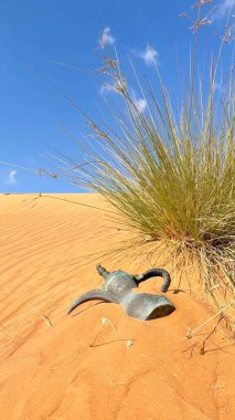 Desert scene features vibrant grass and traditional coffee pot resting on sand. Sand dunes stretch across desert landscape, blending golden sand with clear blue sky in arid beauty. clipart