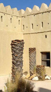 Arap kalesinin avlusunda yüksek palmiye gövdeleri ve doğal çöl manzaraları ön plana çıkıyor. Avlu tasarımı, kale duvarlarını küçük açıklıklarla vurguluyor, çöl işlevselliğini kültür mirasıyla harmanlıyor.