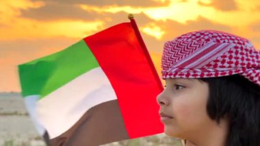 Abu Dabi, BAE - 7 Aralık 2024: Emirati çocuğu miras ve birliği simgeleyen BAE bayrağını gururla taşıyor. Emirati çocuğu ayakta duruyor, ulusal gururunu yansıtıyor, gün batımına karşı bayrak sallıyor. 