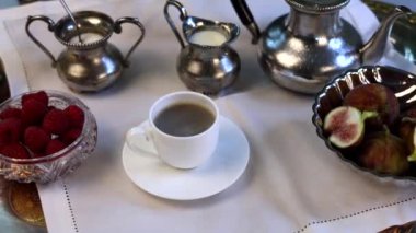 Gümüş çay setiyle besleyici kahvaltı, eşleşen bacaklar, pürüzsüz porselen fincanlara dökülen çay, dekoratif gümüş tepside dinlenen incirler ve kristal kasedeki canlı ahududular..