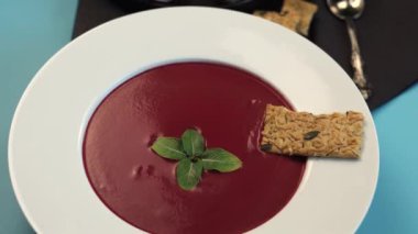 Pırıl pırıl beyaz tabakta servis edilen pancar çorbası taze nane şekeri, kraker, minimalizm, temiz tasarım, kalın görseller, estetik basitlik ile süslenmiş, çarpıcı bir şekilde mavi arka planda sunuluyor..