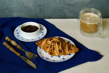 Dekoratif mavi ve beyaz tabakta çikolatalı bir kruvasan ile şeffaf bir çift duvarlı fincanda bir fincan kahveyle eşleştirilmiş bir kahvaltı sahnesi..