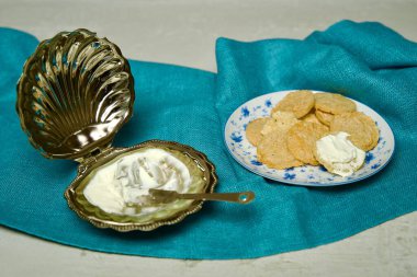 Seramik bir tabakta sebze gevrekleri ve gümüş tereyağı kabında krem peynirden oluşan bir kompozisyon, açık dokulu bir yüzey üzerinde turkuaz bir kumaş üzerinde düzenlenmiştir..