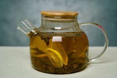 Şeffaf cam bir çaydanlığın yakın plan çekimi. Sert demlenmiş yeşil çay ve hafif desenli bir yüzeye yerleştirilmiş taze limon dilimleri..