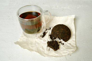 Açık ambalajda siyah Pu-erh çayı yanında şeffaf bir fincan demlenmiş çay ile çayın koyu ve zengin rengini gösterir..