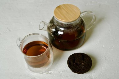 Hafif dokulu bir yüzeye yerleştirilmiş siyah Pu-erh çayı, yanında şeffaf demlenmiş çaydanlık, çayın zengin, koyu rengini vurguluyor..