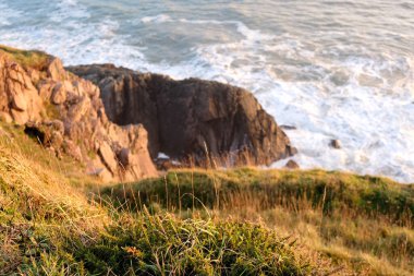  Gün batımında kayalık bir sahil şeridi görüntüsü, ön planda çimenler var.