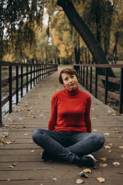 Sonbahar yapraklarıyla çevrili bir parkta oturan olgun bir kadının neşeli bir portresi. O parlak kırmızı bir balıkçı yaka kazak giyiyor. Arka planda altın yapraklı ağaçlar var, rahat ve doğal atmosfere ekleniyor.. 