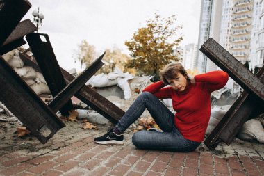 Kırmızı kazaklı bir kadın şehir ortamında büyük bir metal kirişe yaslanmış şekilde yerde oturuyor. Sahne, güç ve şehir yaşamının elementlerini sonbahar ağaçları ve etrafındaki yapraklarla birleştiriyor. 