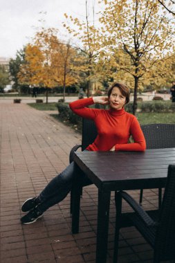 Parkta oturan olgun bir kadının neşeli bir portresi. Parlak kırmızı bir balıkçı yaka kazak giyiyor ve büyük sarı bir yaprak tutuyor. Titreyen sonbahar sezonunun tadını çıkarırken sıcak bir gülümsemeyle.. 