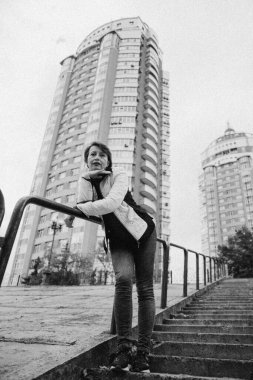 Sıradan bir ceket ve kırmızı balıkçı yaka giyen kendinden emin orta yaşlı bir kadın, arka planda modern yüksek binalar olan bir şehir ortamında düşünceli bir şekilde poz veriyor..