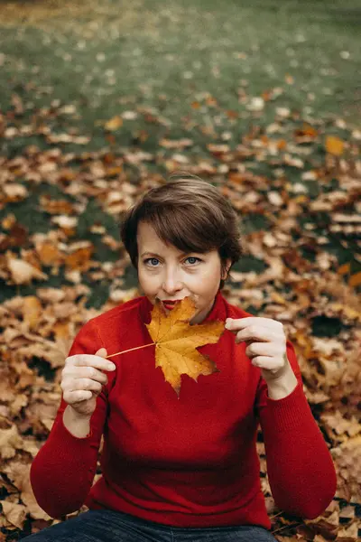 Sonbahar yapraklarıyla çevrili bir parkta oturan olgun bir kadının neşeli bir portresi. Bu fotoğraf sonbaharın güzelliğini yakalar ve doğanın basit zevkleri için mutluluk, rahatlama ve takdir hissini iletir.. 