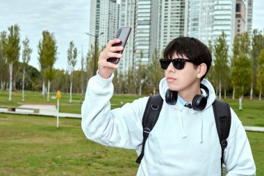 Şehir parkında kulaklıklar ve kapüşonlu kapüşonlu genç adam selfie çekiyor.