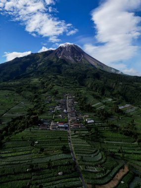 Dikey İHA görüntüsü, Stabelan Köyü 'ndeki evler tarımla çevrili. Görkemli Merapi Dağı, ince dumanlar ve mavi gökyüzü saçan tepe noktasıyla güzel bir zemin sağlar..