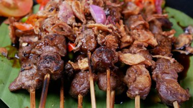 Soya soslu yumuşak keçi satay, kızarmış arpacık soğanı serpiştirilmiş, taze domates sambal, limon dilimleri ve siyah tabakta domates ile servis edilmiş..