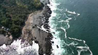 Karang Taraje plajında, Sawarna, Banten, Endonezya 'da, yoğun bir ormanın arka planına sert dalgaların çarptığı kayalık bir sahilde insansız hava aracı hareketi.