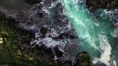 Havadan bakıldığında okyanus dalgaları siyah kayalara çarparak koyu yeşil deniz suyuyla çelişen beyaz köpük oluşturur. Su akışı güçlü görünüyor, engebeli kaya yarıklarını aşıyor..