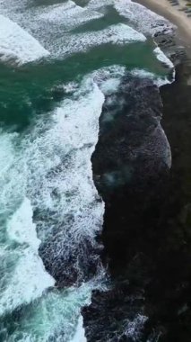 Sert dalgaların sert bir şekilde siyah kayalara çarptığı kayalık bir sahilin üzerindeki hava manzarası güzel ama çok tehlikeli görünüyor. Banten, Endonezya Sawarna plajı..