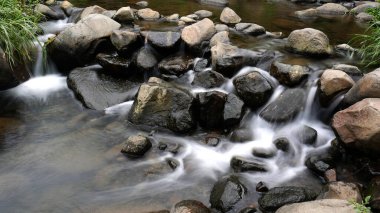 Nehir, büyük doğal kayalar arasında akar ve yeşil yapraklarla nehir kenarlarını süsleyen sakin bir manzara yaratır..
