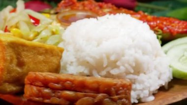 Baharatlı kırmızı sambal, kızarmış tofu, kızarmış tempeh, taze sotelenmiş sebze, karides ezmesi sambal, salatalık dilimleri ve diğer taze kenarlarla servis edilen bir tabak sıcak beyaz pirinci kapat..