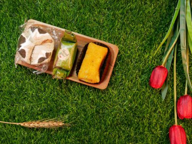 Kırmızı çiçekler, buğday sapları ve sentetik çim bazlı tatlı ve lezzetli Bolu Kukus, Risoles ve Lemper içeren geleneksel Endonezya pasta paketi..