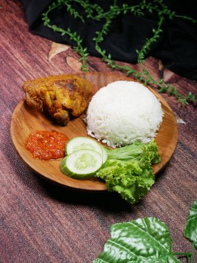 Geleneksel Endonezya yemekleri beyaz pirinç, kızarmış tavuk, baharatlı kırmızı Sambal, taze salatalık dilimleri ve yuvarlak ahşap bir tabakta marul yapraklarından oluşur. Yeşil plastik yapraklarla süslenmiş..