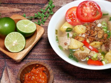 Baharatlı kırmızı biber sosu ve taze limonla servis edilen et çorbası, patates, domates ve kızarmış arpacık soğanı. Kahverengi arka planda beyaz bir kasede servis edilmiş.