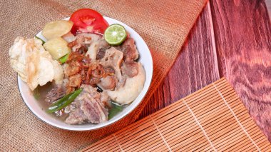 Sop buntut or oxtail soup in a white bowl with fresh potatoes, tomatoes, lime and fried onions. Served in a traditional style on a bamboo mat and burlap. Space for text available clipart