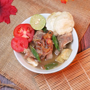 Bir kase sığır pirzolası çorbası ya da biftek kemiği, patates, taze domates, limon dilimleri ve kızarmış arpacık soğanı. Bambu paspaslar, çuval bezleri ve yapraklarla servis edilir.