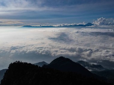Tepenin siluetinin ve tepesindeki ağaçların havadan görünüşü. Arkaplan dramatik bir bulut denizi. Uzakta Bromo Tengger Semeru Ulusal Parkı görkemli bir şekilde görülür..