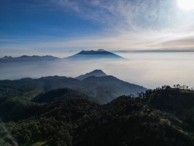 İnsansız hava aracının hava manzarası yeşil tepelerin ve otlakların doğal güzelliğini ön planda gösteriyor. Arka planda, Arjuno Dağı Welirang kuleleri ince bir sabah sisi ile örtülüydü..