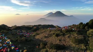 Sakin ve huzurlu atmosfer şafak vakti Prau Dağı 'nda görülür. Dağcıların renkli çadırları, altın yeşili otlaklar ve diğer dağlar ince sabah sisi ile kaplı olarak yükselirken görülür..