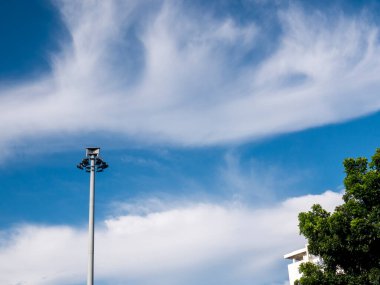 Üzerinde yeşil ağaçlar ve binalarla çevrili sokak lambası olan metalden yapılmış uzun sokak lambası direği. Arka plan olarak inanılmaz bulutlu mavi gökyüzü. Sokak lambası halkın güvenliği için çok yararlıdır..