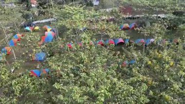 Hava manzarası, Dronlar dağlardaki bir kamp alanının üzerinde uçuyor. Renkli çadırlar, yemyeşil ağaçların altında düzgünce düzenleniyor. Sabah güneşi serin ve sakin bir kamp hissi veriyor..