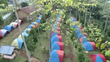 Uçan dronlar açık doğadaki kamp alanının güzelliğini gösteriyor. Mavi ve kırmızı çadırları düzgün bir şekilde dizilmiş, yeşil ağaçlarla çevrili, soğuk ve sakin bir kamp ortamı sağlıyor..