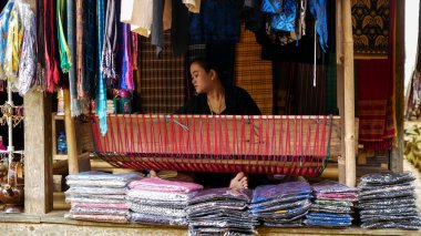 Banten, Endonezya, 15 Kasım 2023. Dış Baduy kabilesinden bir kadın Baduy bambu evinde geleneksel dokuma tezgahı kullanıyor. Etrafında, renkli dokuma kumaşlar alıcıları bekliyor..