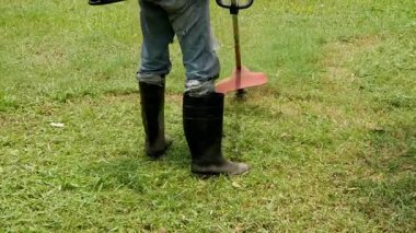 Bir adam, çim biçme makinesi ve güvenlik ayakkabısı kullanan, uzun otlarla kaplanmış bir bahçedeki çimleri kesmeye çalışıyor..