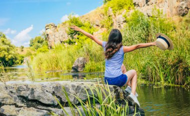 Şapkalı, esmer, genç bir kadının arka plan fotoğrafı. Oturup nehire bakıyor ve dinlenirken eğleniyor..