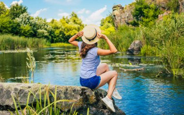 Şapkalı, esmer, genç bir kadının arka plan fotoğrafı. Oturup nehire bakıyor ve dinlenirken eğleniyor..
